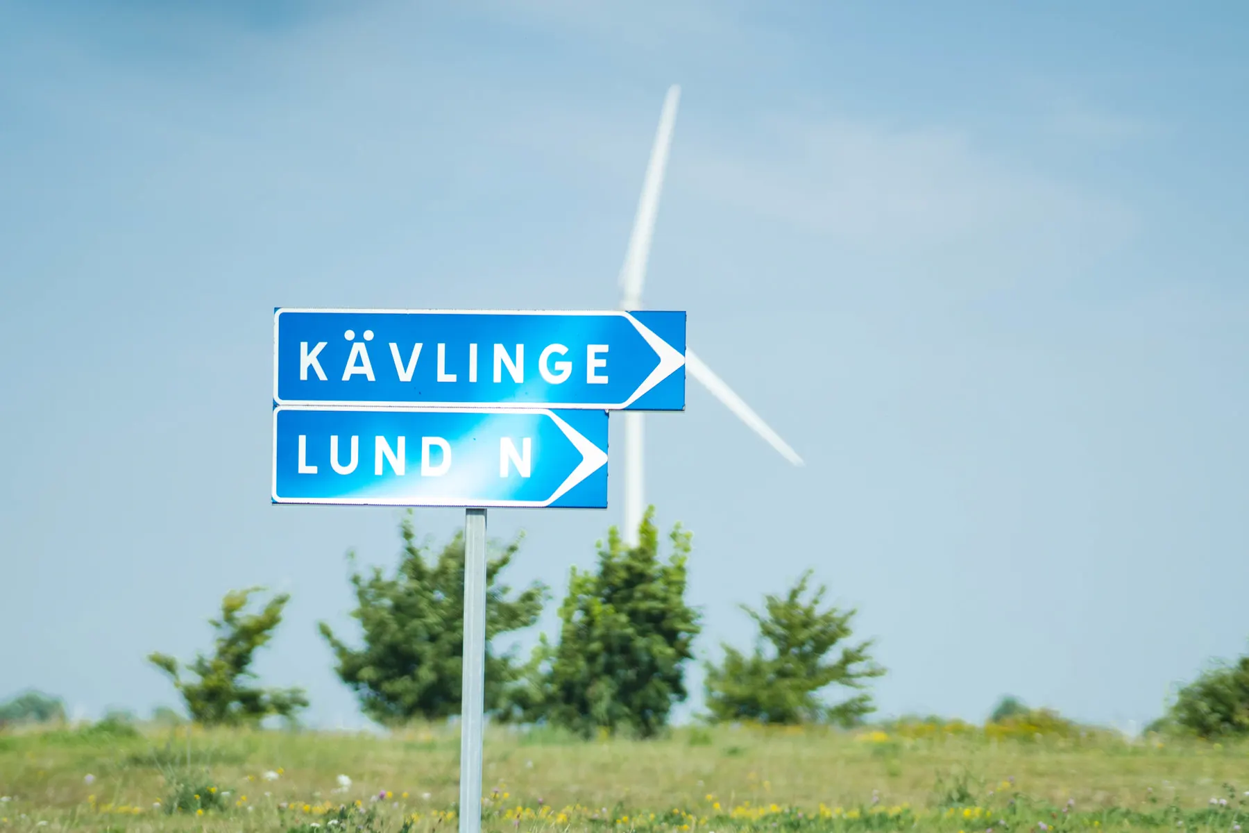 Blå vägvisare mot Kävlinge och Lund N på en stolpe i skånskt landskap med ett vindkraftverk i bakgrunden